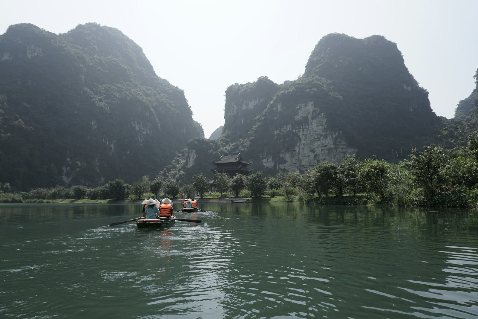 Tourists in life vests on rowboats glide through water flanked by dramatic limestone karsts and lush greenery in Ninh Bình, Vietnam.