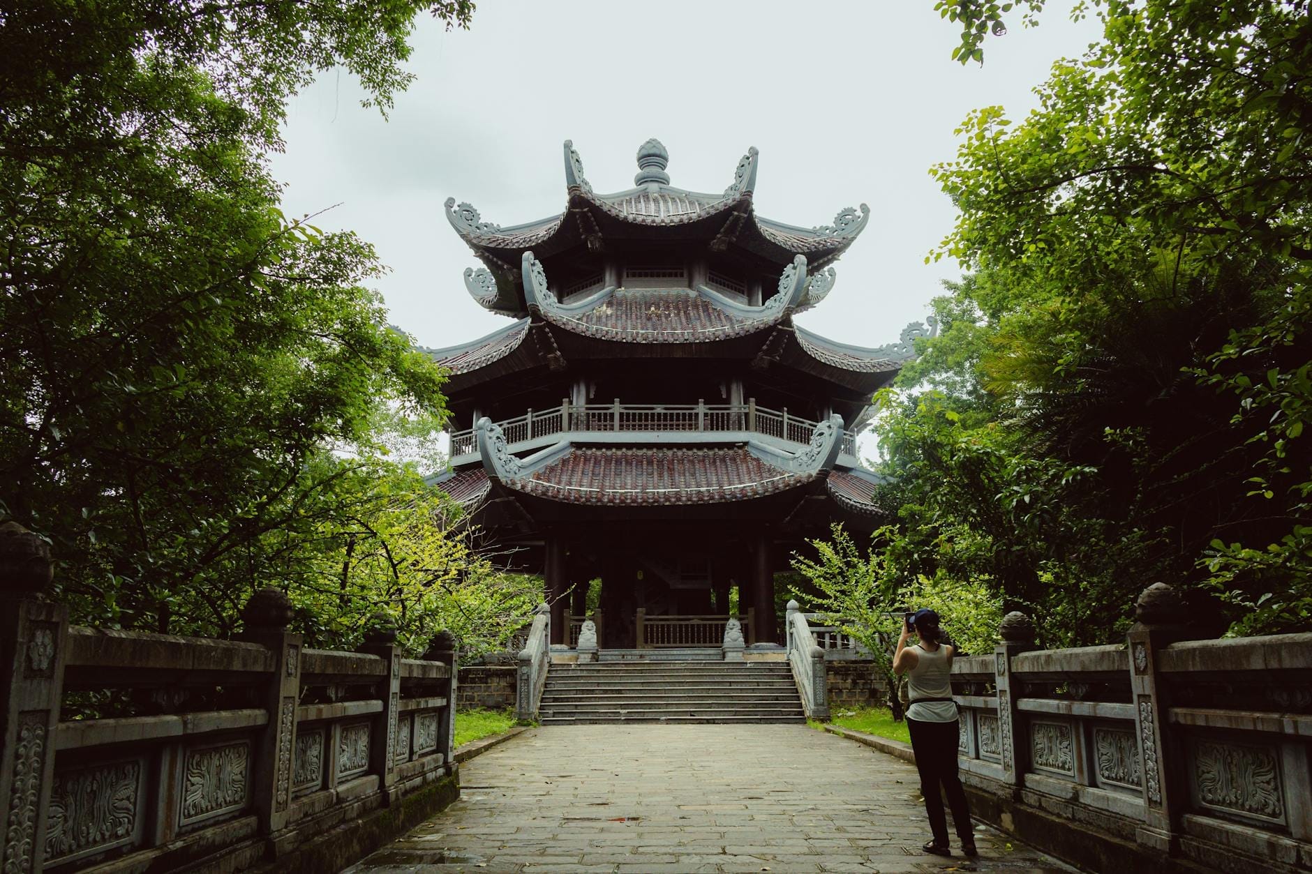 Traditional multi-tiered Buddhist pagoda surrounded by lush greenery, with a visitor walking along a stone pathway leading to the temple.