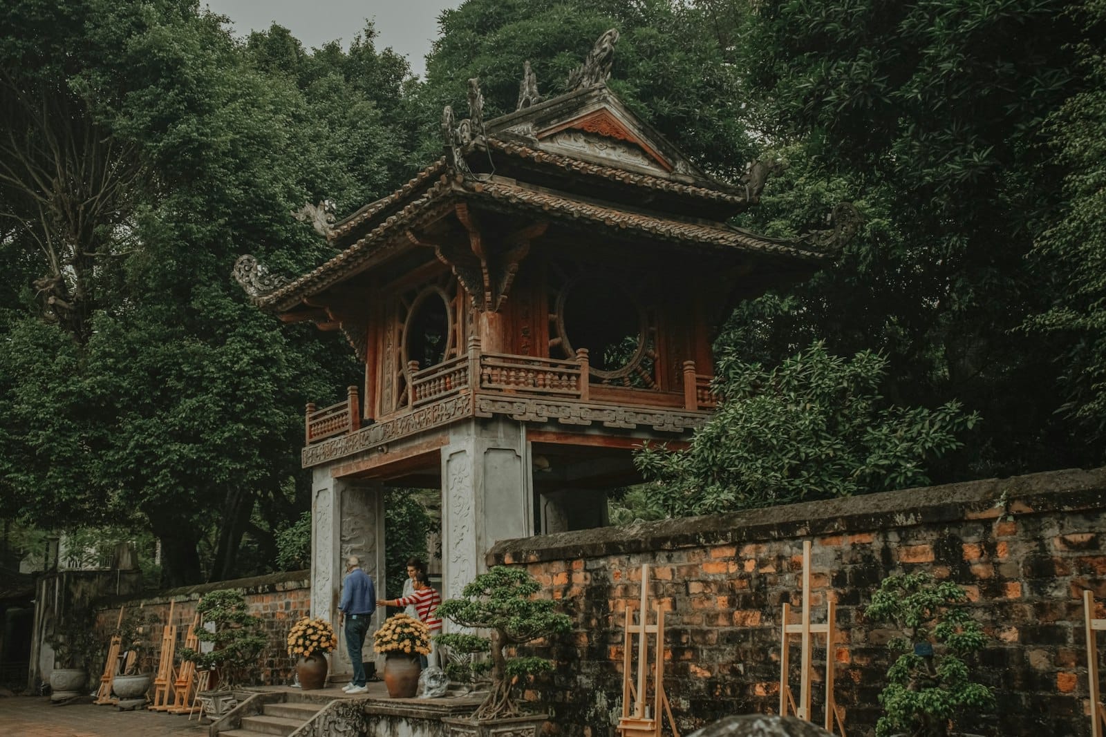 Traditional Vietnamese temple gate surrounded by lush trees and brick walls, with two visitors standing underneath the archway.