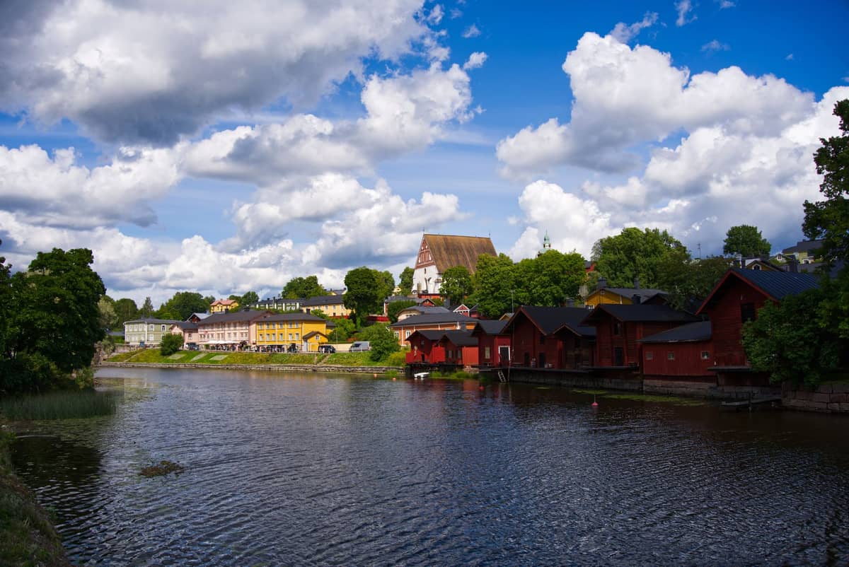 Vista clássica da cidade antiga de Porvoo com armazéns vermelhos à beira do rio, casas de madeira coloridas e uma igreja sob um céu azul com nuvens.