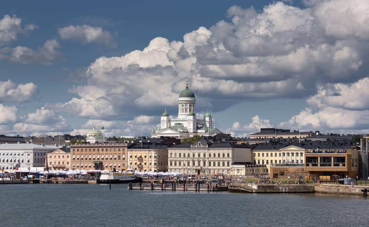 Vista ampla do orla de Helsinque com prédios históricos coloridos, porto movimentado e a Catedral de Helsinque sob nuvens dramáticas ao fundo.