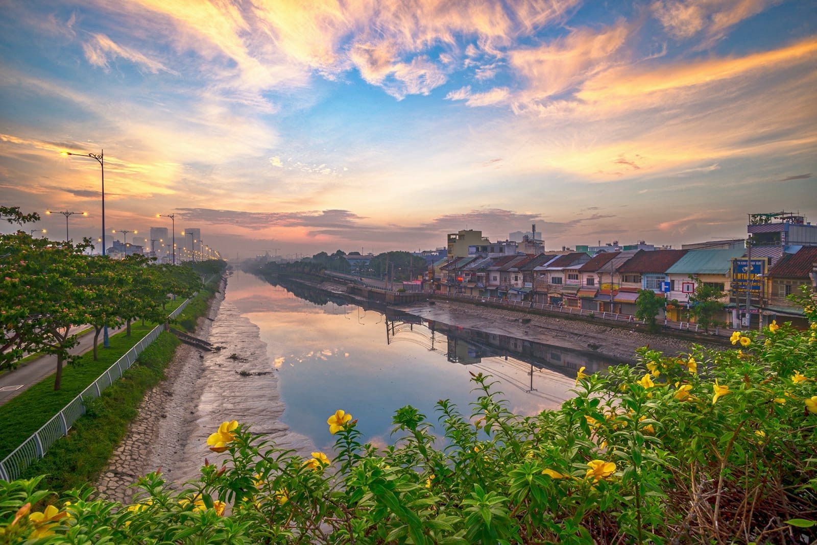 Canal fluvial colorido al amanecer con plantas en flor en primer plano y casas a orillas del agua en Ciudad Ho Chi Minh, una invitación para explorar.