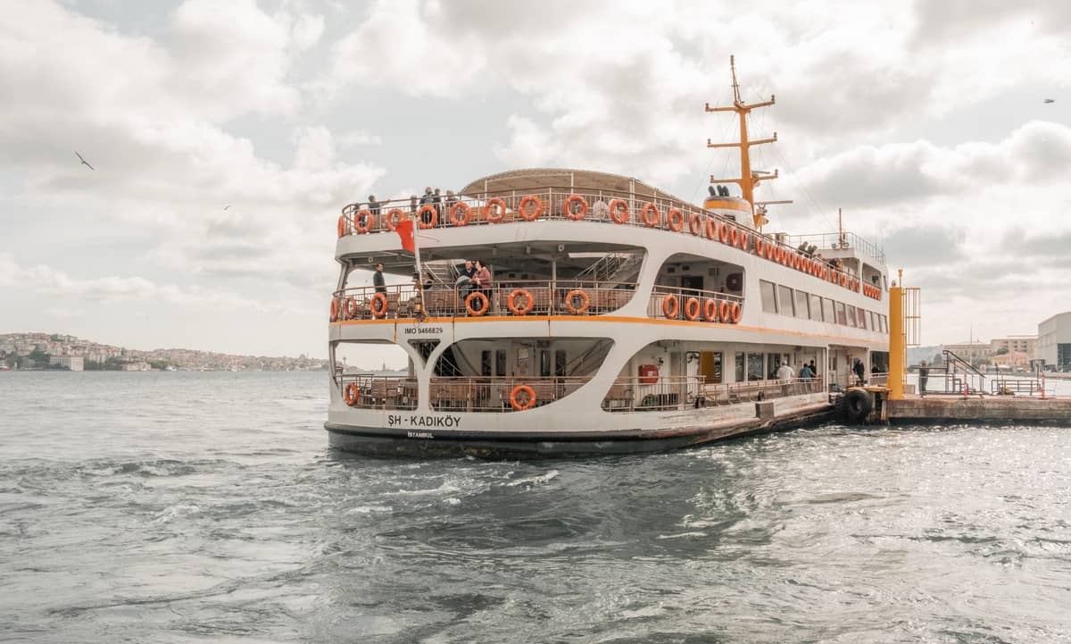 İskelede yanaşmış, güvertesinde yolcuların göründüğü ve arka planda şehir kıyısının yer aldığı 'Kadıköy' yazılı büyük bir İstanbul vapuru.