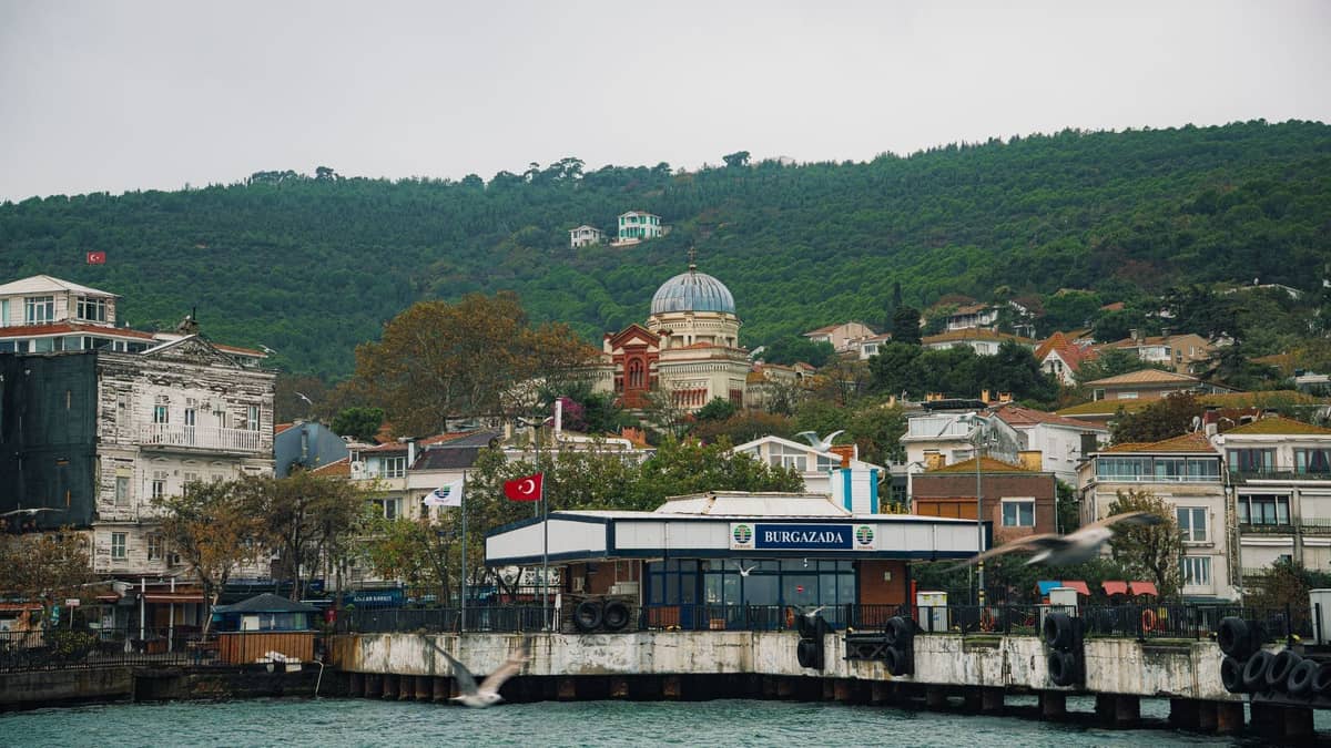 Adalar'da tarihi binalar, çam ağaçlarıyla kaplı tepeler ve kubbeli bir kilise ile çevrili Burgazada iskele terminalinin deniz cephesinden görünümü.