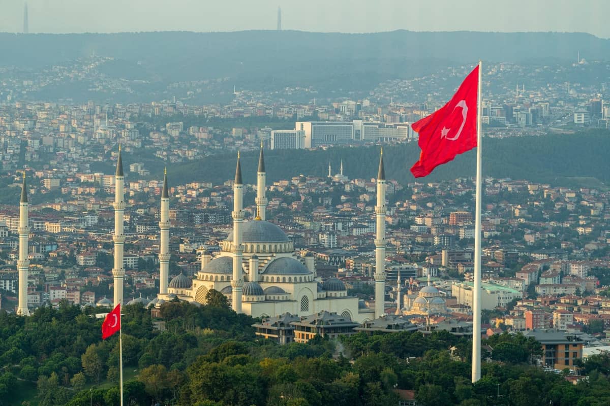 Tepe üzerindeki Çamlıca Camii, Türk bayrakları ve kentsel mahalleler ile uzak tepelerin geniş manzarasıyla İstanbul'un panoramik görünümü.