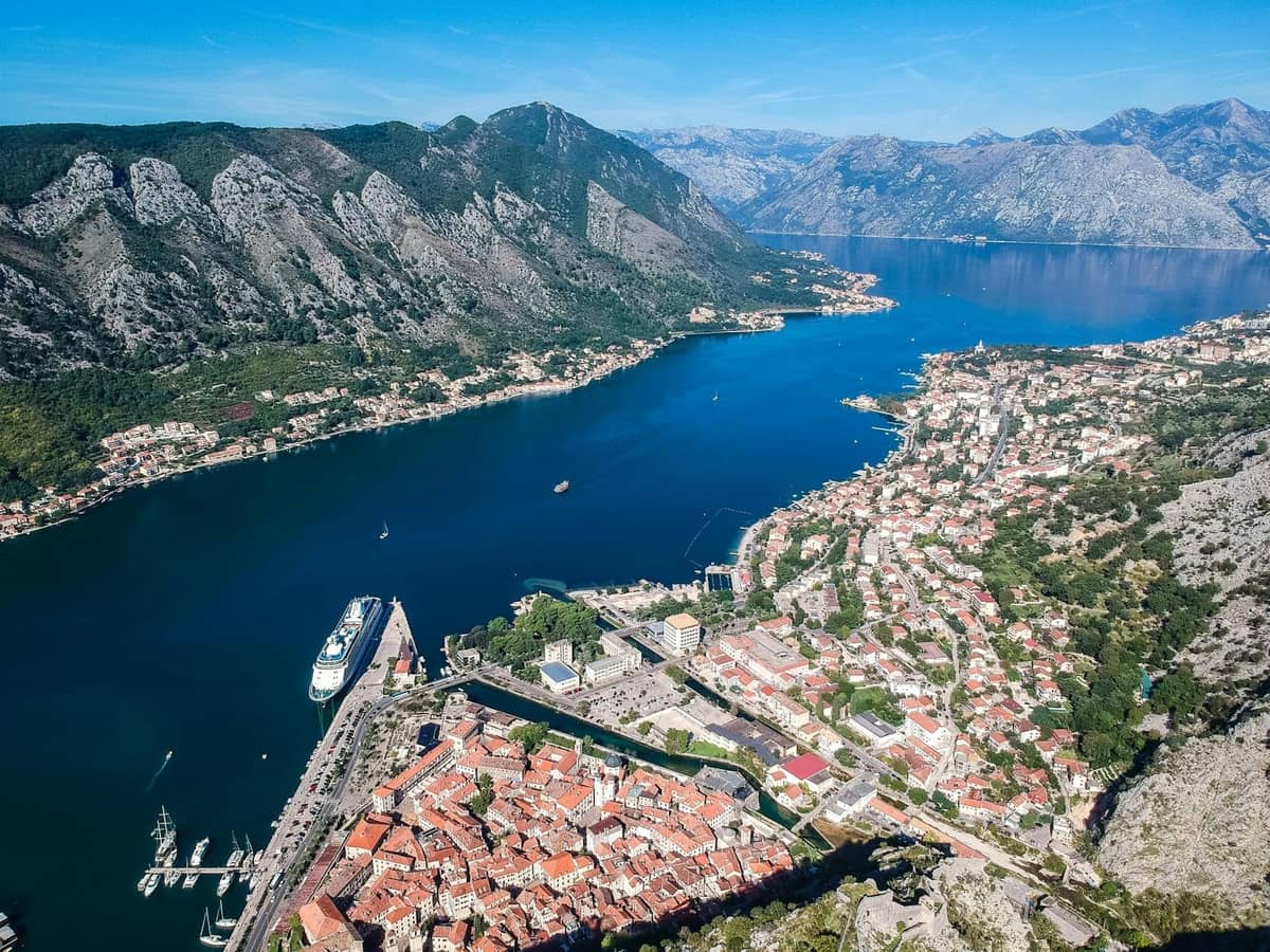 Luftaufnahme von Kotor mit der Altstadt mit roten Dächern, einem an der Bucht angedockten Kreuzfahrtschiff, den umliegenden Bergen und einer weitläufigen Küstenlandschaft unter klarem blauem Himmel.
