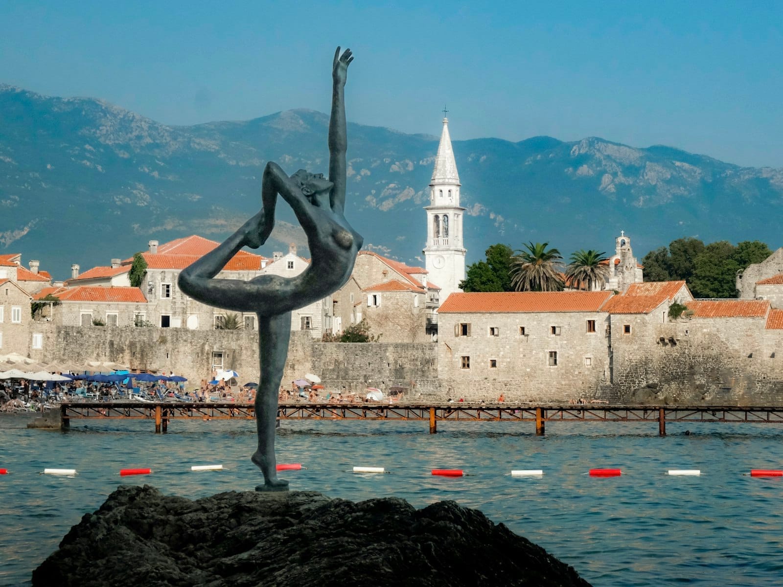 A icônica estátua da bailarina junto ao Mar Adriático com os edifícios de pedra e a torre da igreja da Cidade Velha de Budva tendo as montanhas ao fundo.