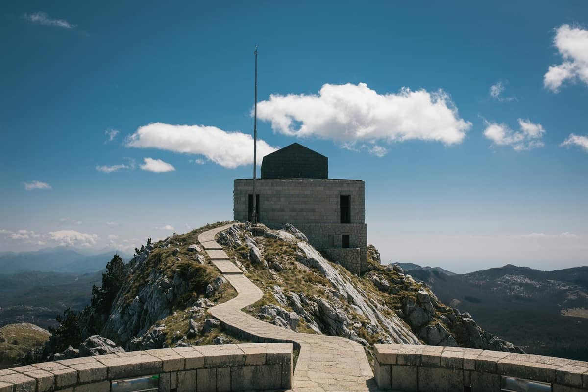 Steinpfad führt einen felsigen Bergrücken zum Njegoš-Mausoleum im Nationalpark Lovćen hinauf, mit Panoramablick auf die Berge unter blauem Himmel.