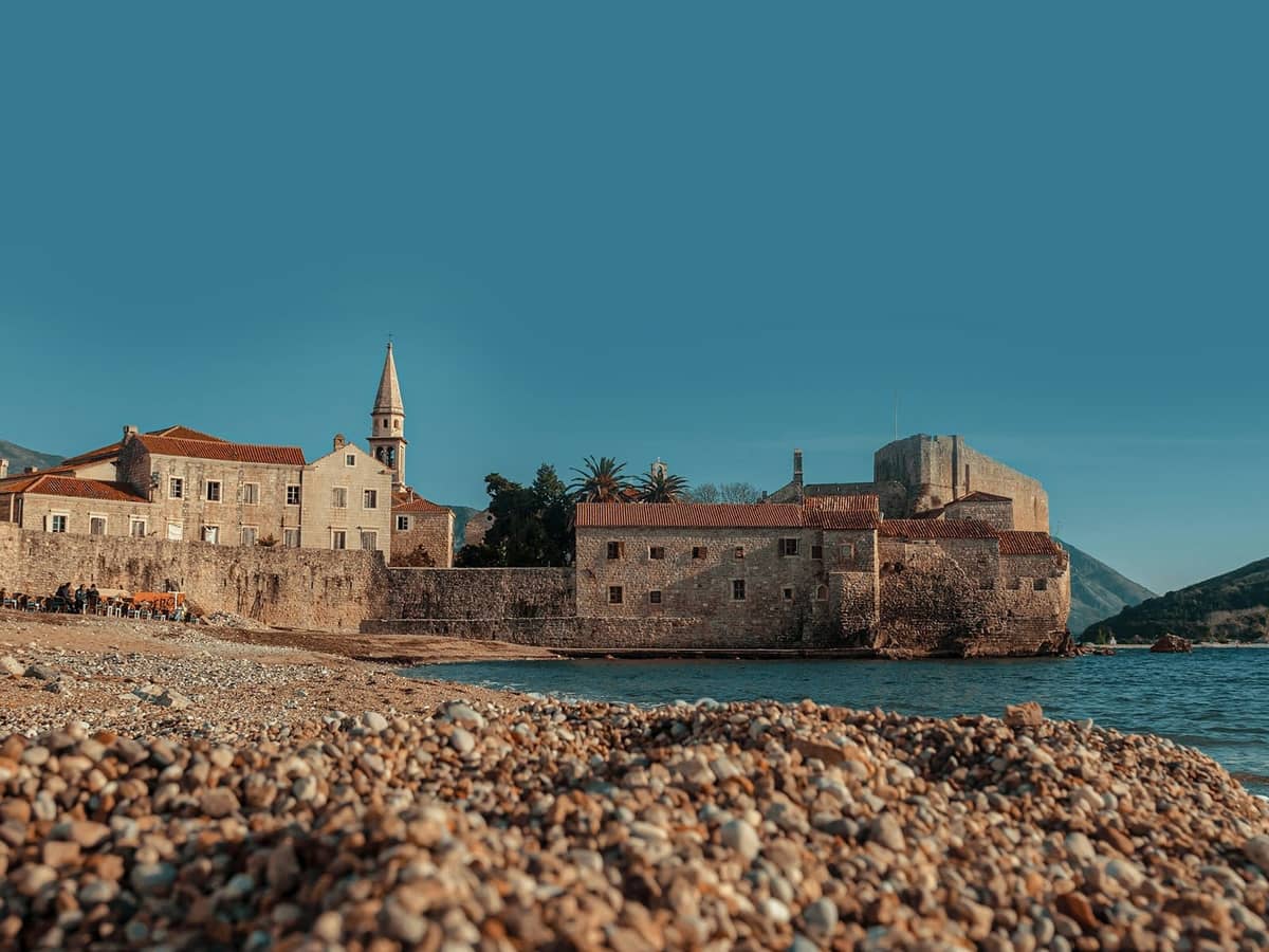 Steinummauerte Altstadt von Budva mit Kirchturm und Festung, von einem Kieselstrand unter klarem blauem Himmel aus gesehen.