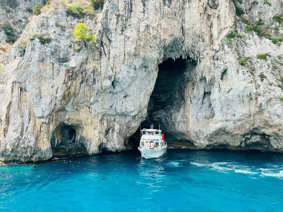 Ein weißes Ausflugsboot mit Passagieren an Bord nähert sich dem Eingang einer felsigen Meereshöhle mit leuchtendem blauem Wasser.