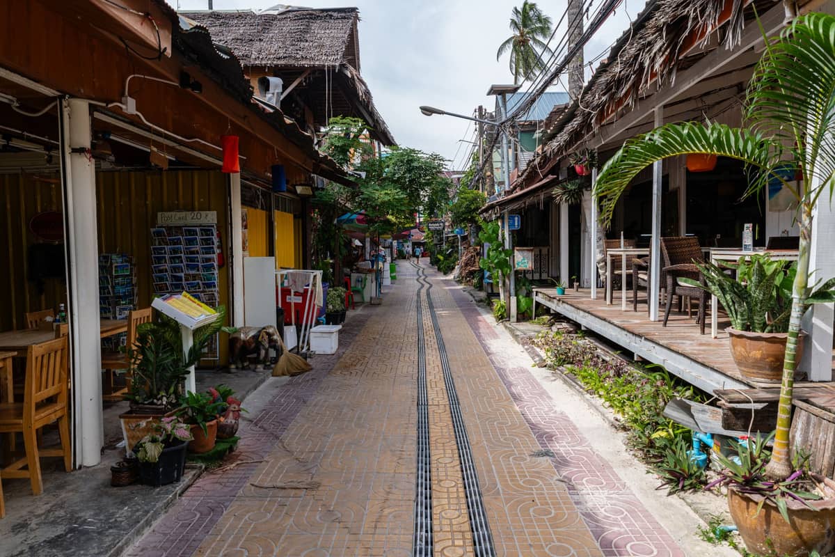 Calle peatonal estrecha en la Tailandia tropical, bordeada de pequeños cafés locales, tiendas y plantas frondosas, típica del pueblo de la isla Koh Lanta.