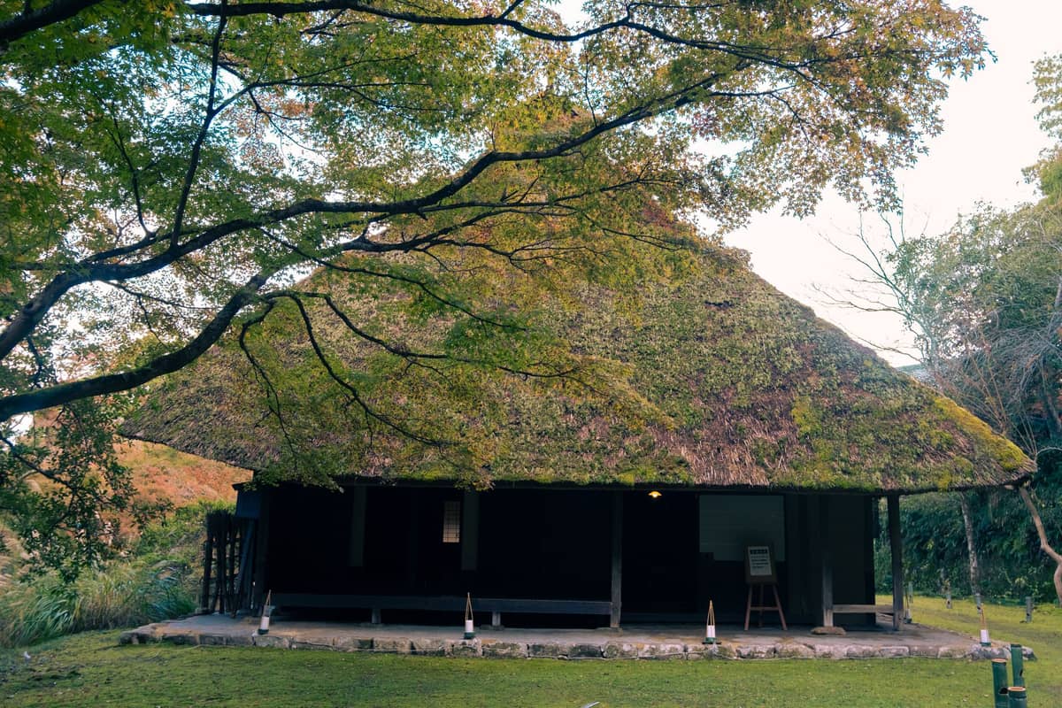 Traditional Japanese farmhouse with a thick thatched roof, surrounded by greenery and trees in a rural, peaceful valley setting.