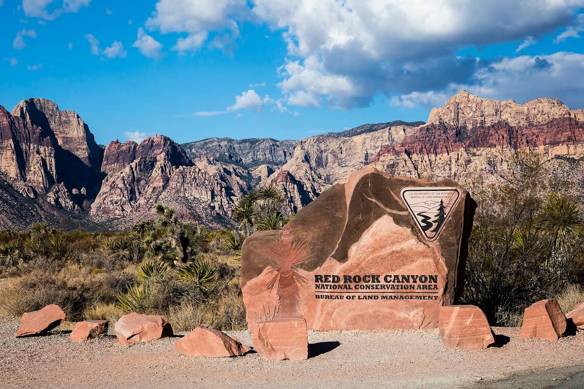 Placa de entrada da Red Rock Canyon National Conservation Area com dramáticas montanhas de rocha vermelha e paisagem desértica ao fundo sob um céu azul brilhante.