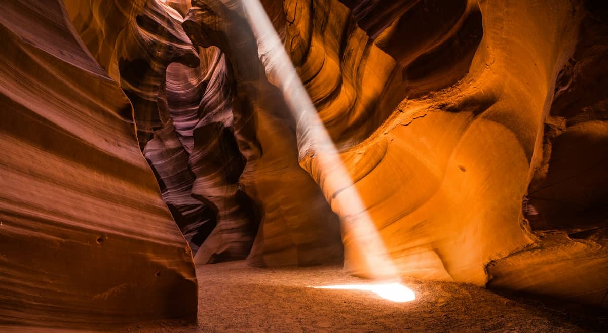 Vista panorâmica do interior do Antelope Canyon com impressionantes paredes de rocha laranja e um dramático feixe de luz solar iluminando o chão do canyon.
