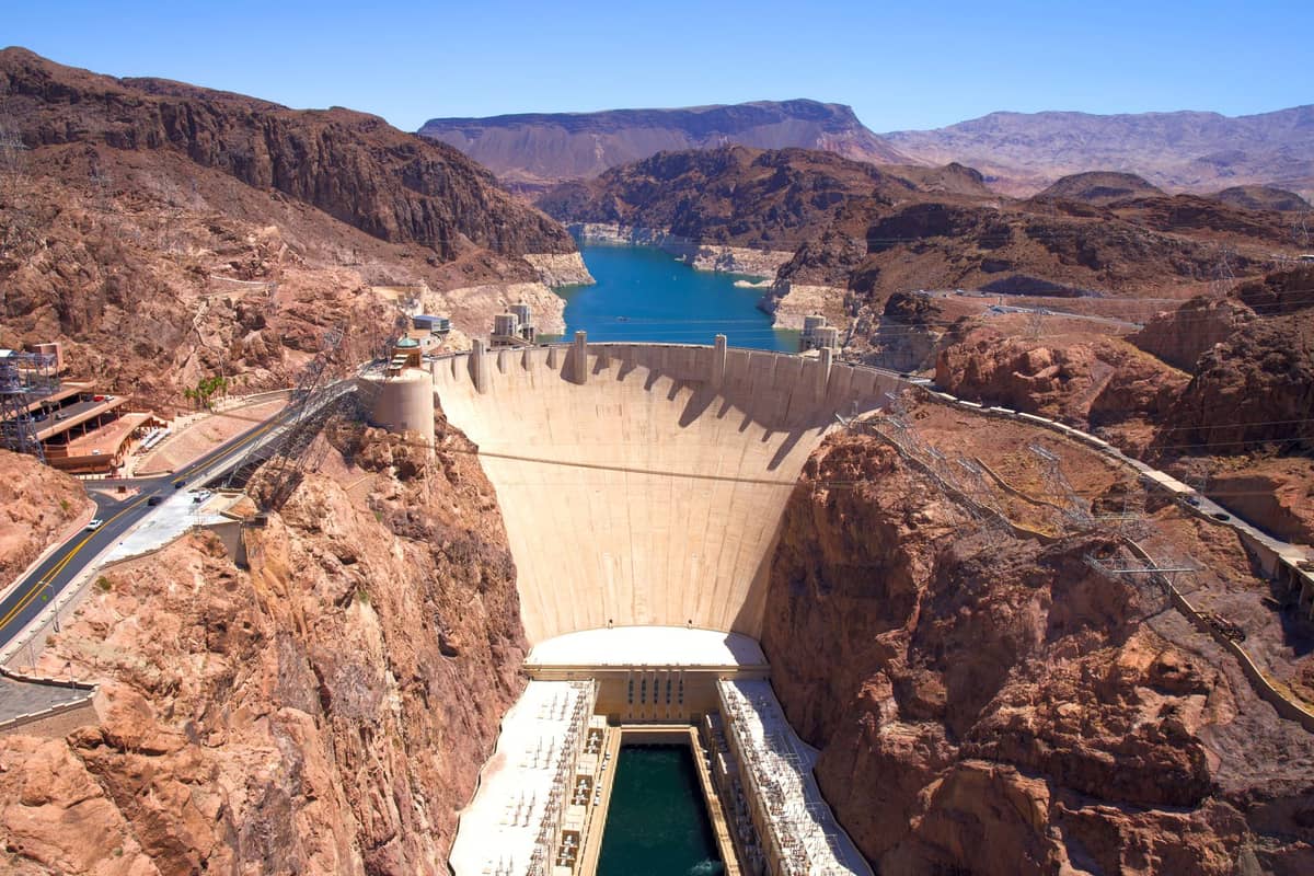 Vista aérea ampla da Hoover Dam situada entre colinas rochosas do deserto, com água azul brilhante se estendendo ao longe sob um céu limpo.