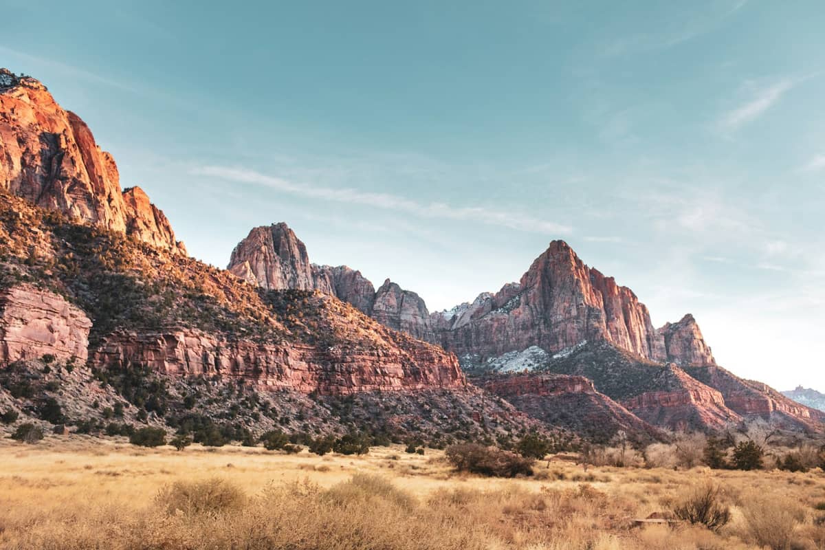 Vista ampla de picos montanhosos avermelhados dramáticos e campos amarelos sob um céu azul, típicos do Zion National Park perto de Las Vegas.