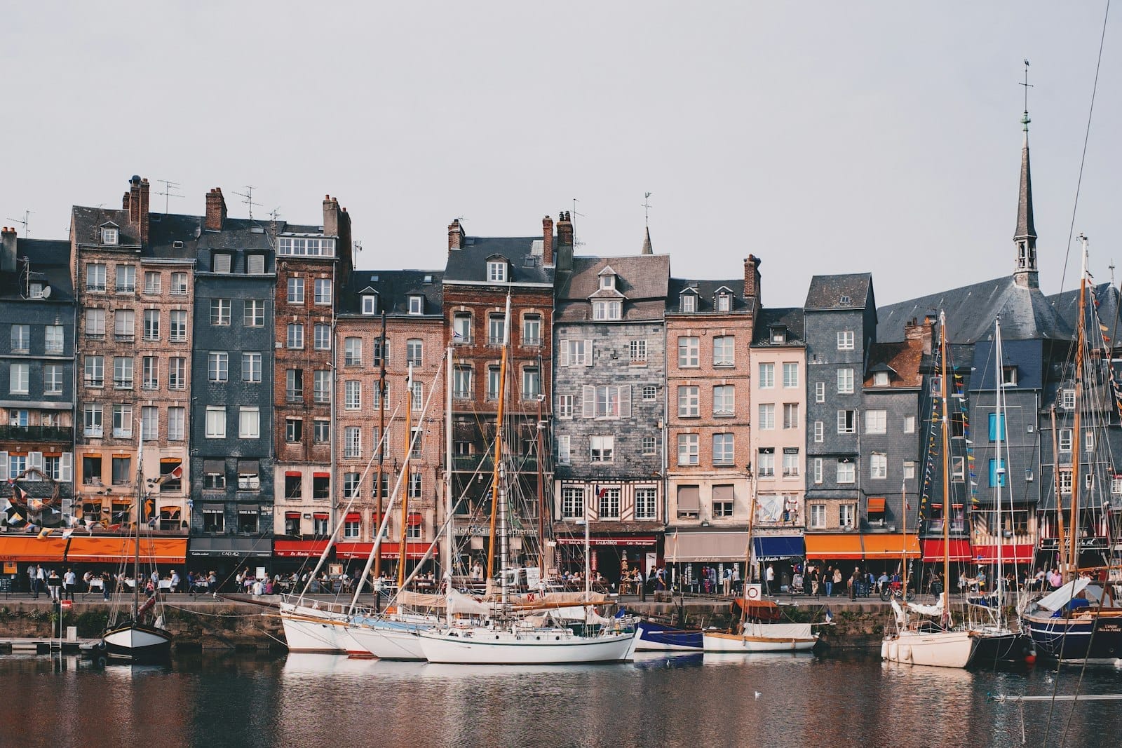 Rumah-rumah tinggi berwarna-warni yang berjajar di sepanjang pelabuhan bersejarah Vieux Bassin di Honfleur, dengan perahu berlabuh dan pantulannya di air.
