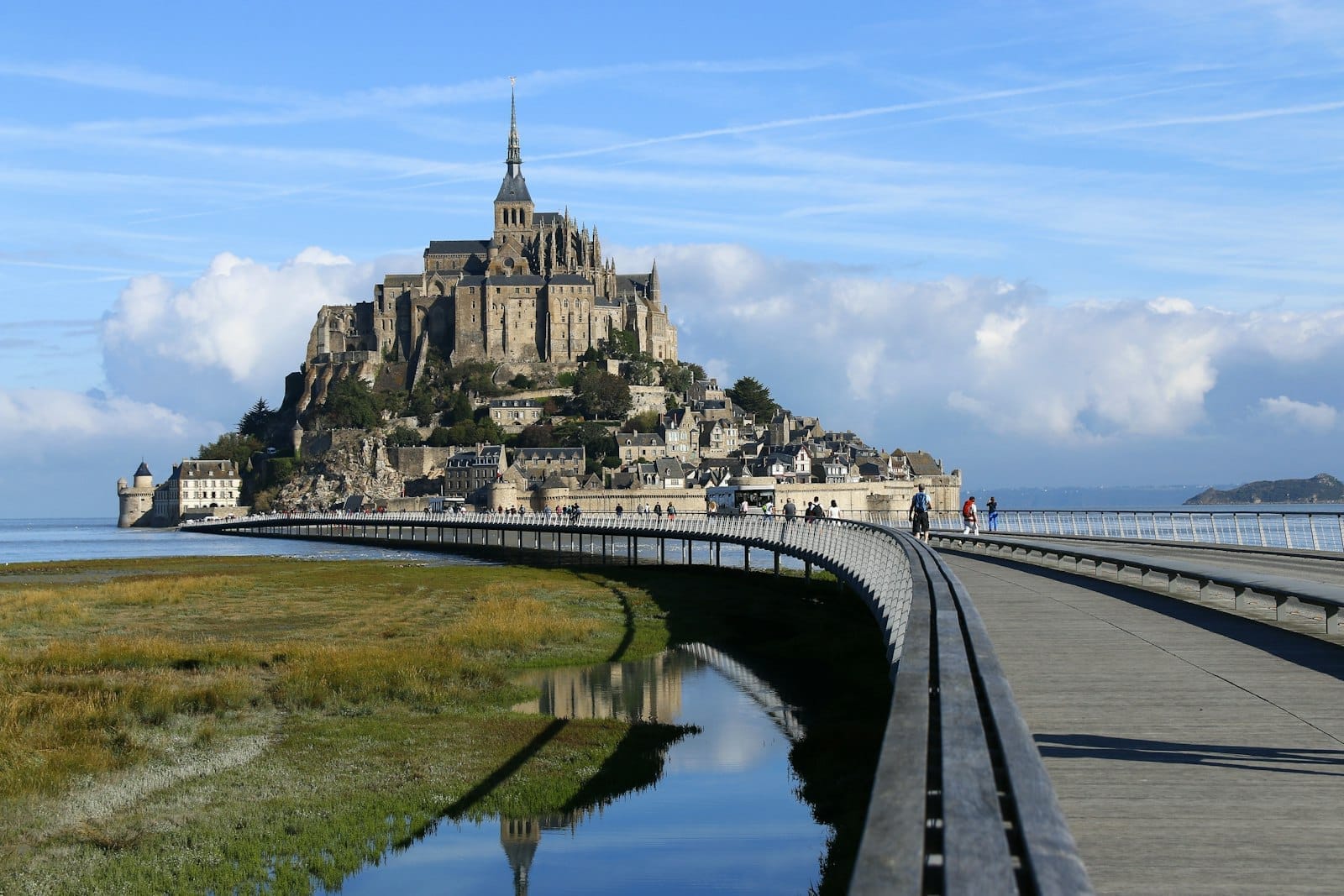 Mont-Saint-Michel dengan jalan lintas yang ditinggikan dan orang-orang berjalan di bawah langit biru, diabadikan dari kejauhan pada hari yang cerah