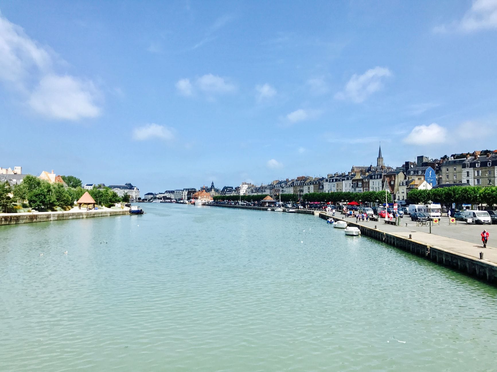 Vista panoramica del porto di Honfleur con i suoi edifici storici, le barche ormeggiate lungo la banchina e un cielo azzurro con qualche nuvola: tutto il fascino costiero della Normandia.