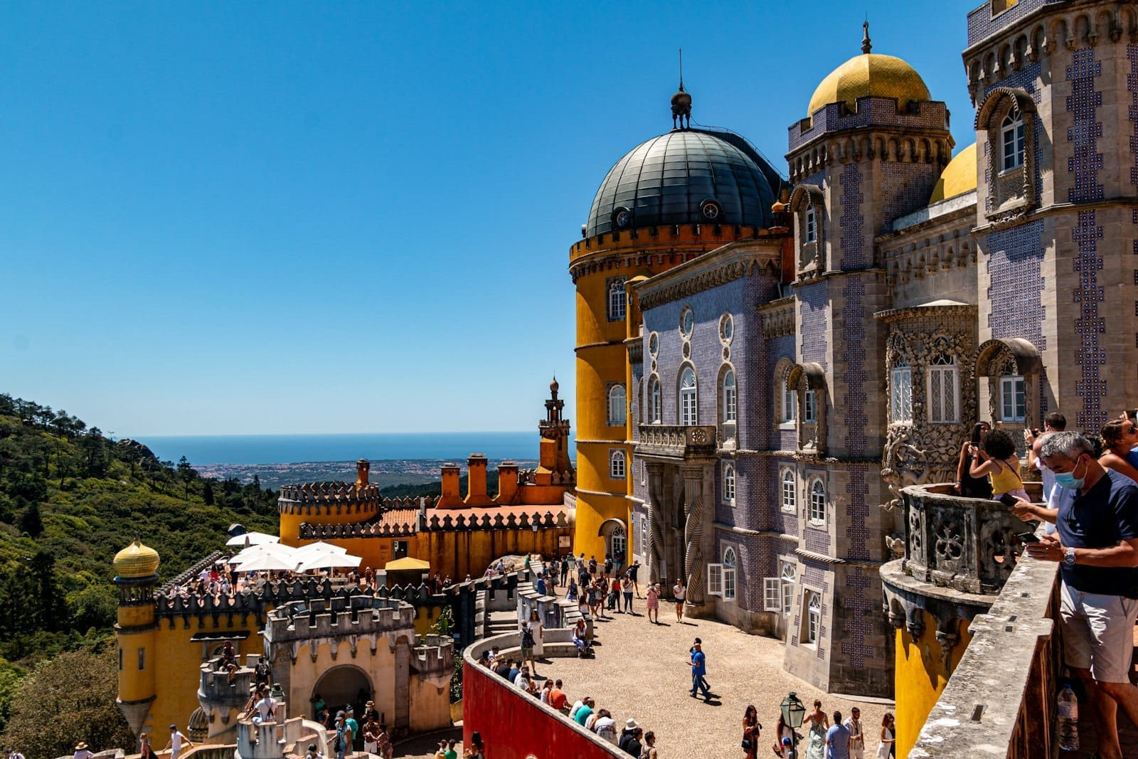 Le coloré Palais de la Pena à Sintra avec des foules de visiteurs, des tours décoratives et des collines verdoyantes sous un ciel bleu.