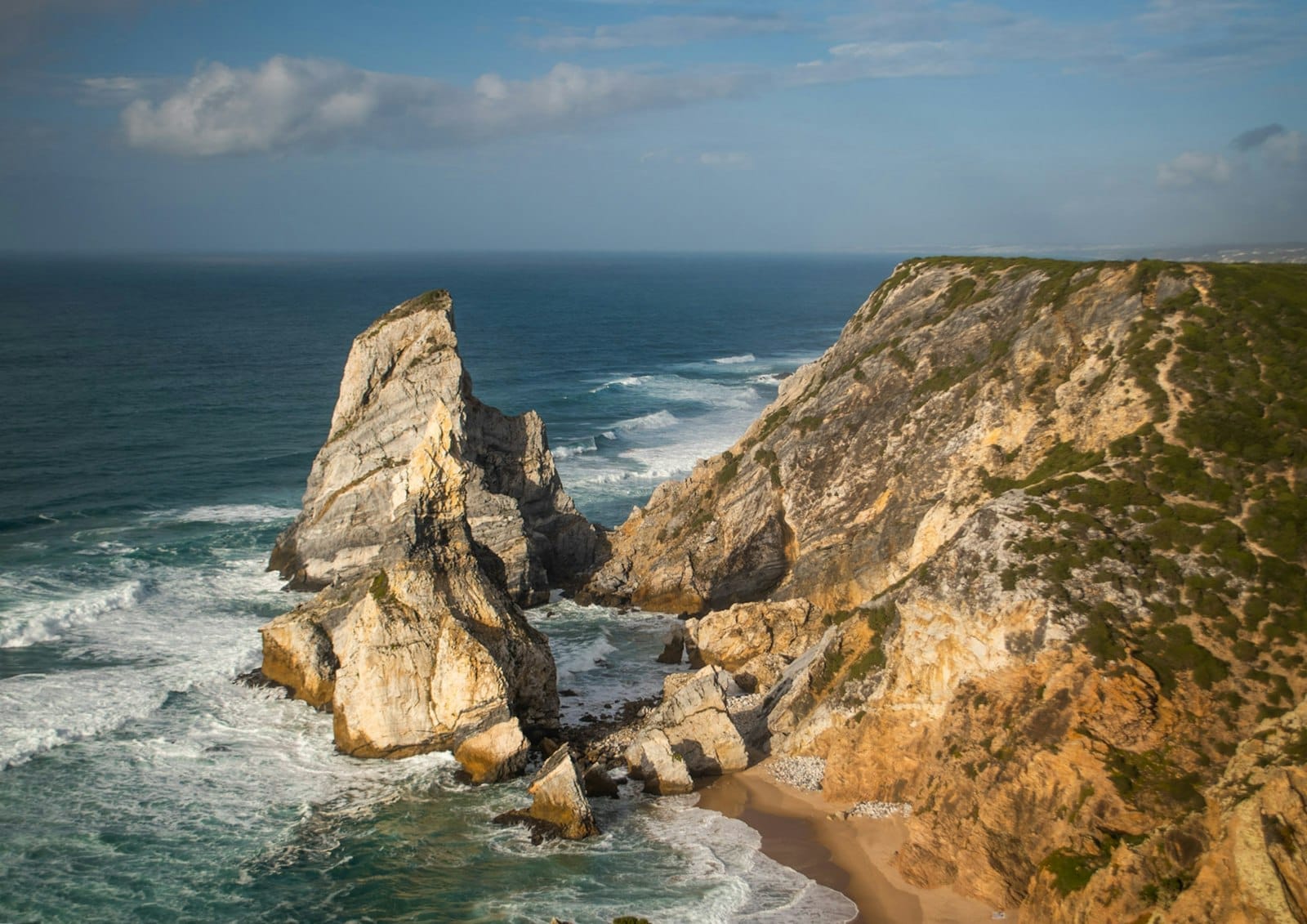 Falaises côtières spectaculaires et grandes formations rocheuses plongeant dans l'Océan Atlantique avec des vagues se brisant en contrebas et un ciel bleu au-dessus.