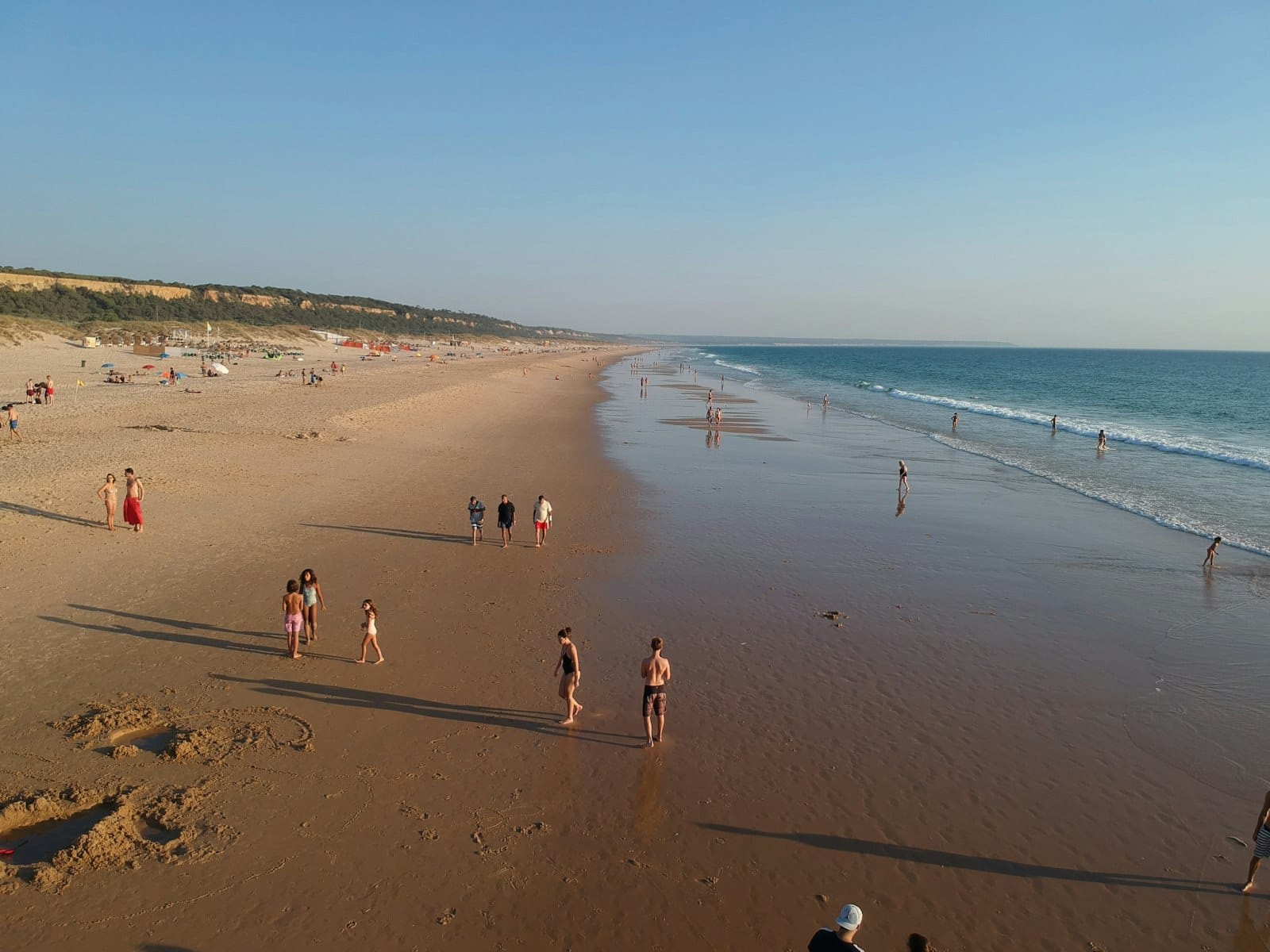 Grande plage de sable avec des personnes se promenant et jouant le long du rivage sous un ciel bleu dégagé, typique de Costa da Caparica au sud de Lisbonne.