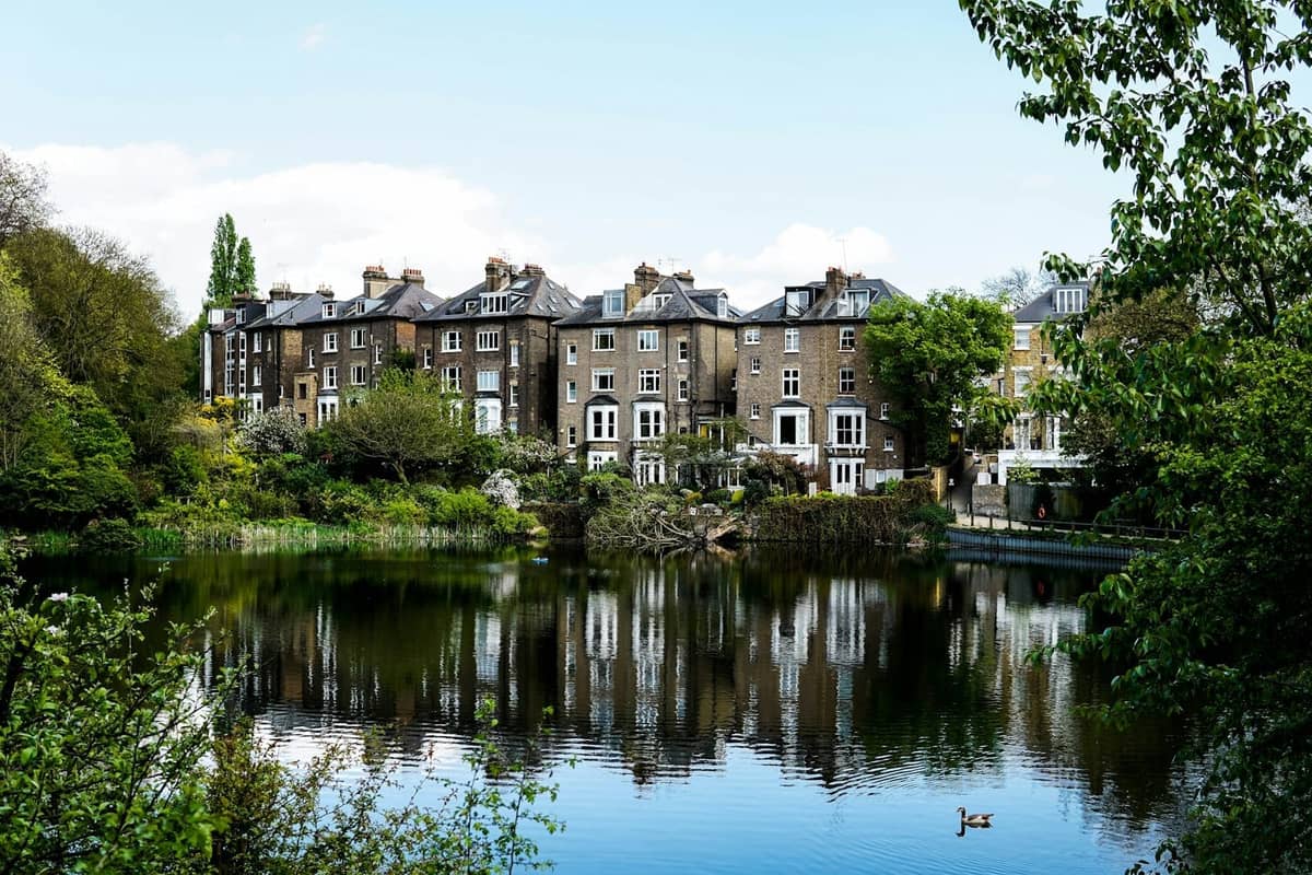 Lago com vegetação em frente a casas geminadas clássicas do norte de Londres, refletindo na água rodeada de árvores, sob um céu luminoso.