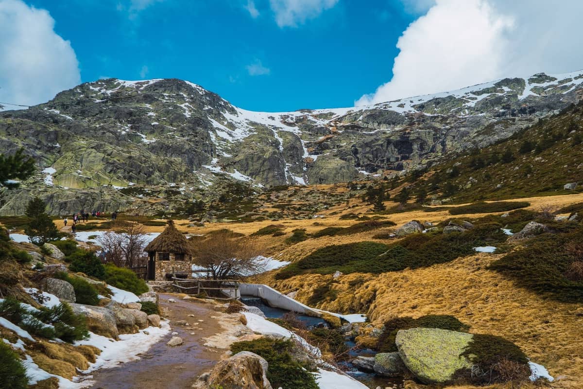 Gebirgslandschaft in der Sierra de Guadarrama mit Schneefeldern, felsigem Gelände, einem Bach und einer kleinen Holzhütte unter einem strahlend blauen Himmel.