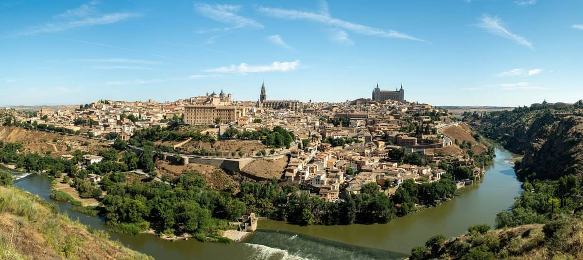 Panoramablick bei Tag auf Toledo, mit der historischen Skyline, dem Fluss und dem berühmten Alcázar prominent im Zentrum, unter einem klaren blauen Himmel.