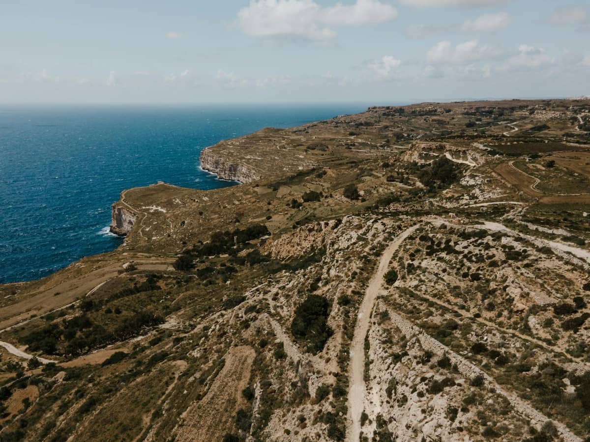 Luftaufnahme von dramatischen Küstenklippen und rauer Landschaft, die unter einem teilweise bewölkten Himmel auf das blaue Mittelmeer treffen.