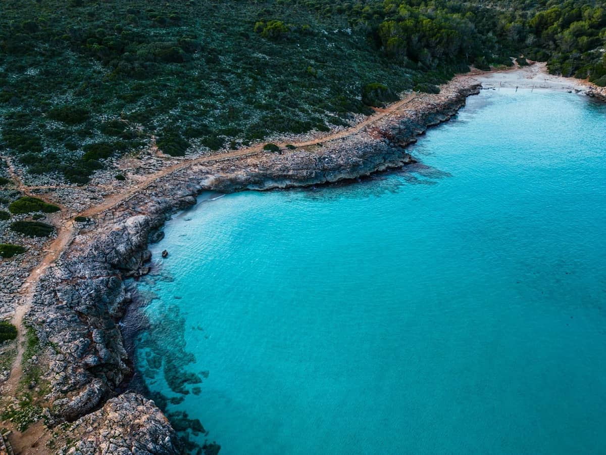 Luftaufnahme einer kleinen felsigen Bucht mit kristallklarem türkisfarbenem Wasser und spärlicher Vegetation, ähnlich der Blue Lagoon auf Comino, Malta.