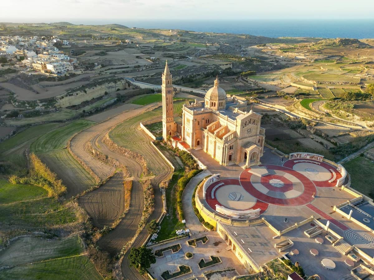 Luftaufnahme der Basilika Ta' Pinu auf der Insel Gozo, umgeben von terrassierten Feldern und dem fernen Meereshorizont.