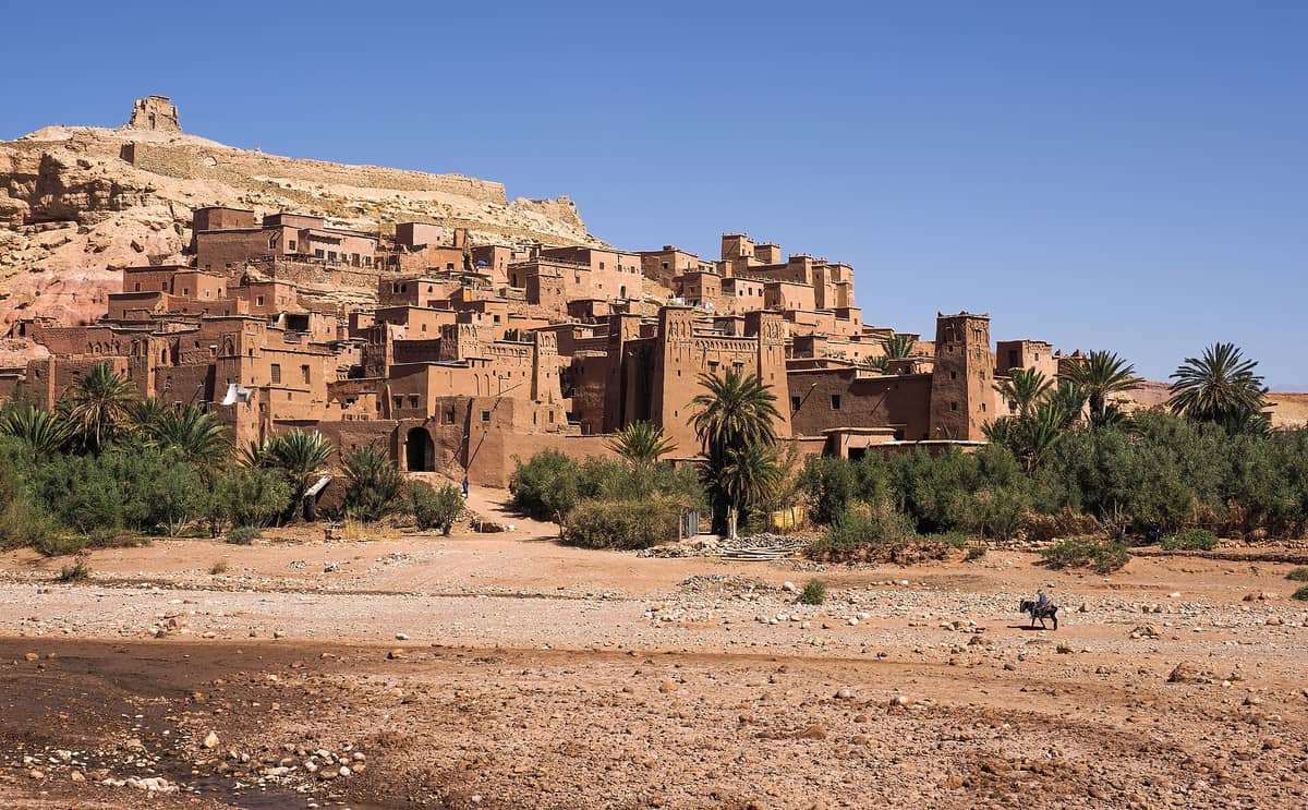 วิวกว้างของ Ait Benhaddou ป้อมคาสบาห์ดินชื่อดังที่มีหอคอยและบ้านเรือนหลายหลัง ล้อมรอบด้วยพืชพรรณเบาบาง ภายใต้ท้องฟ้าสีฟ้าใส