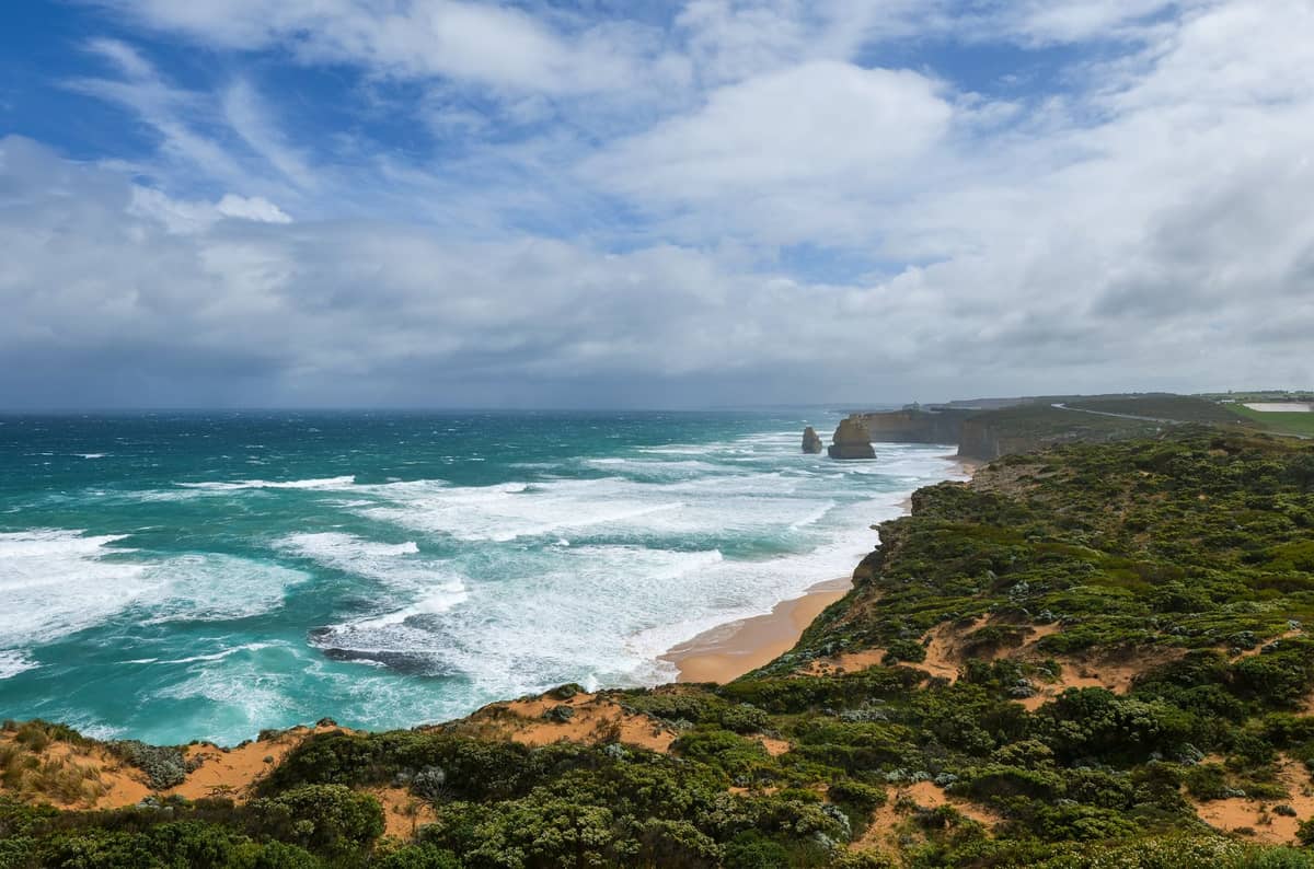 Impresionante costa de la Great Ocean Road con vegetación en los acantilados, olas rompientes y dos columnas de caliza de los Doce Apóstoles visibles en el mar.