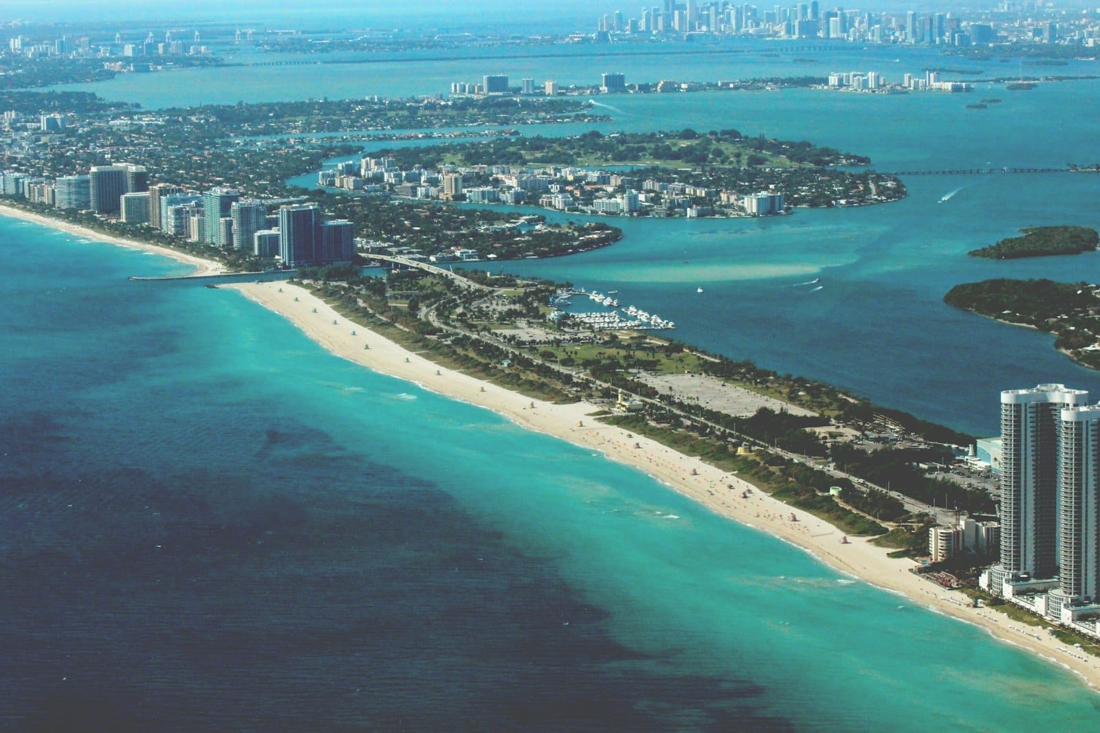 Vista aérea panorâmica mostrando o litoral, as praias e as ilhas perto de Miami, com águas turquesa e o skyline da cidade ao fundo sob um céu azul brilhante.