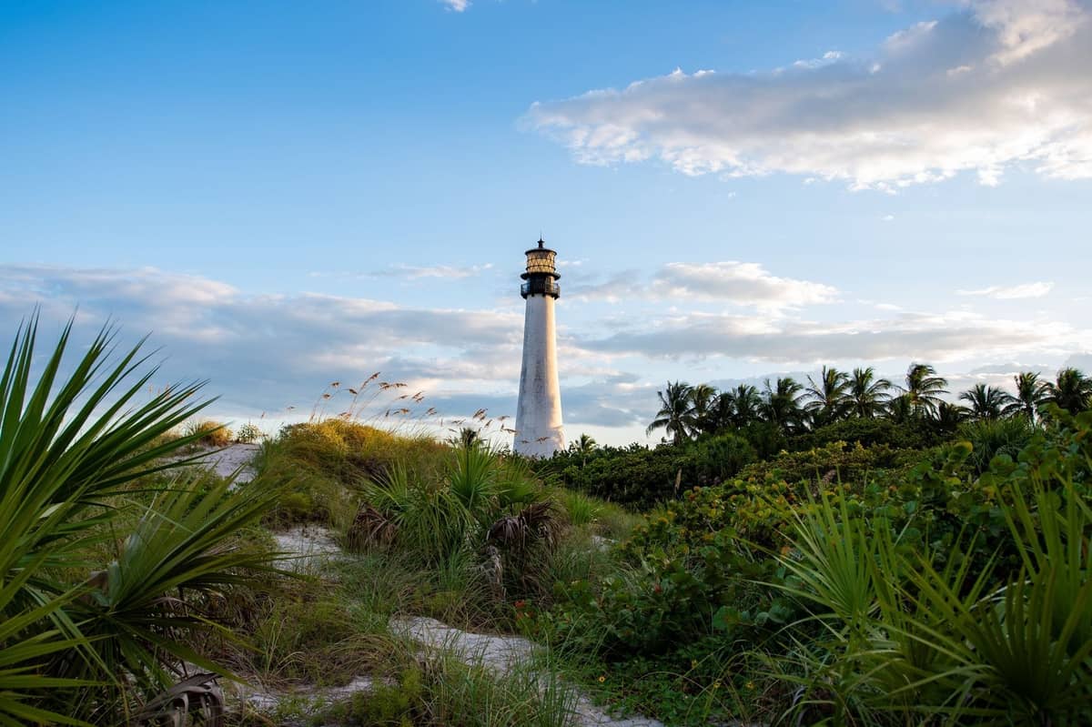 A tall white lighthouse rises above lush tropical greenery and palm trees under a blue sky with scattered clouds.