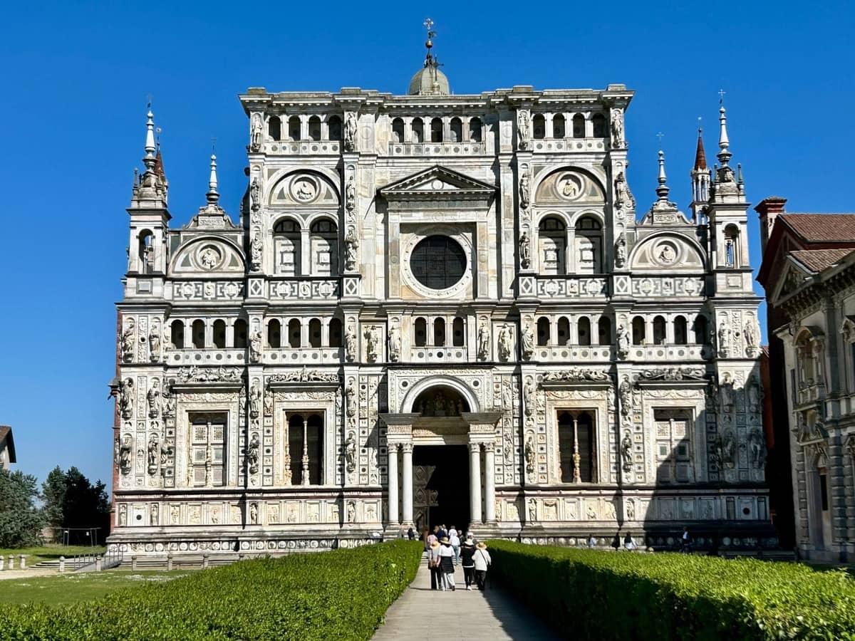 Ornate white marble facade of the Certosa di Pavia monastery with visitors walking towards its entrance on a sunny day