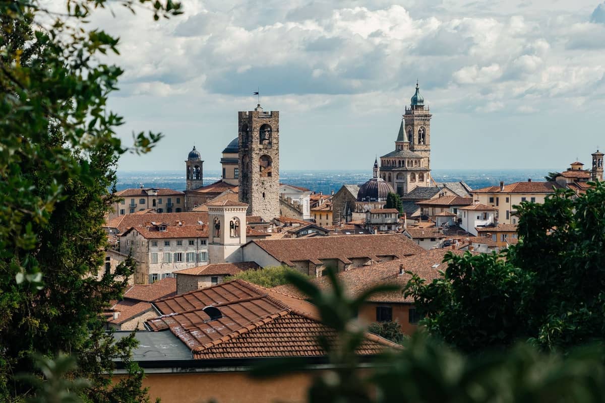 Panoramic view of Bergamo's medieval skyline with historic towers and church spires rising above terracotta rooftops, framed by trees.