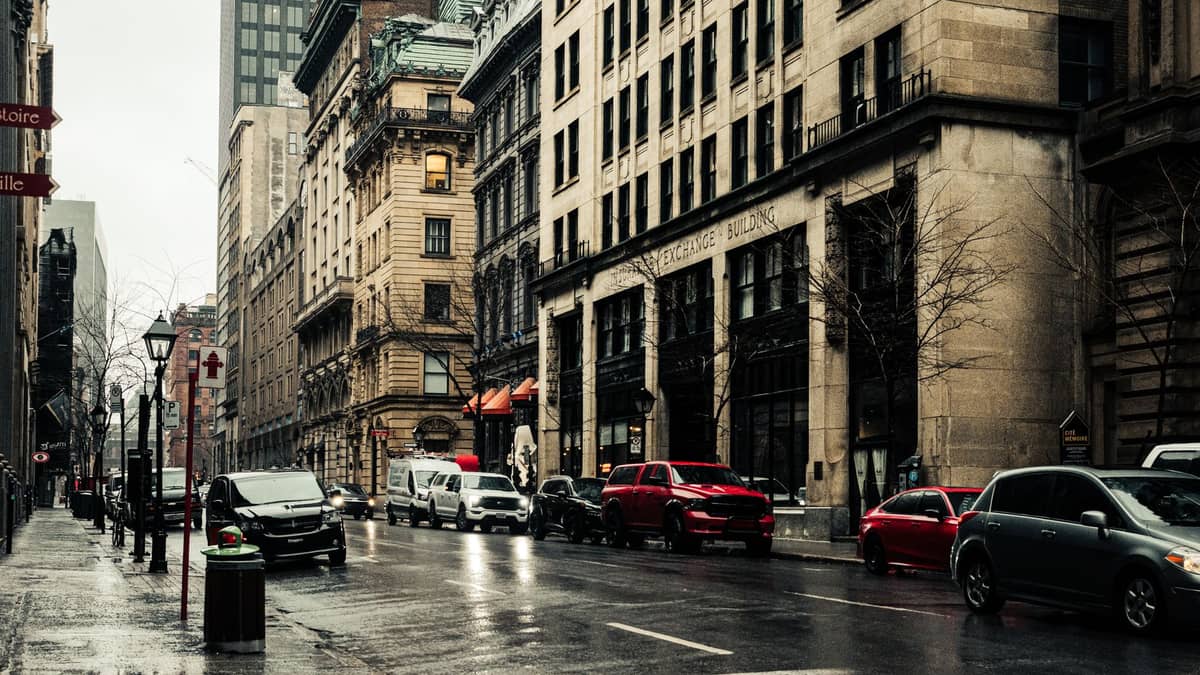Strada del centro di Montreal con edifici storici in pietra, auto parcheggiate lungo il marciapiede e negozi in una giornata nuvolosa.