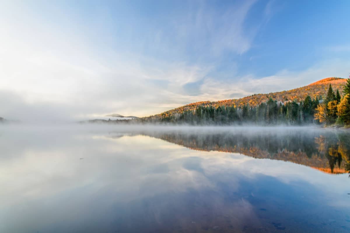 Lago autunnale nebbioso con colline boscose e vivaci colori autunnali riflessi nell'acqua calma sotto un cielo azzurro nelle Laurentides.