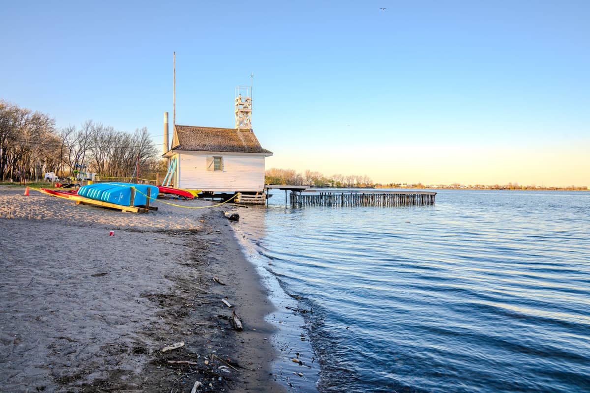 Spiaggia sabbiosa con un capanno per barche sul lago, kayak e acque calme sotto un cielo azzurro limpido vicino a Montreal.