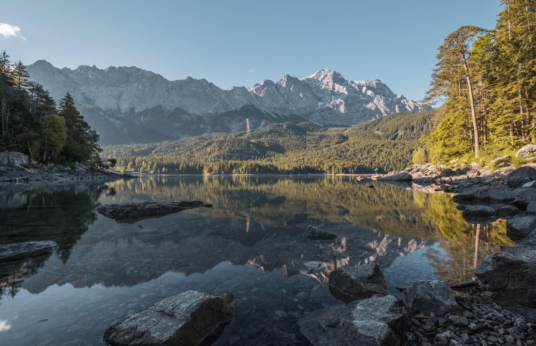 ทะเลสาบใสสะท้อนภาพยอดเขา Zugspitze ที่ปกคลุมด้วยหิมะและยอดเขาโดยรอบ มีป่าและหินอยู่เบื้องหน้าใต้ท้องฟ้าสีฟ้า