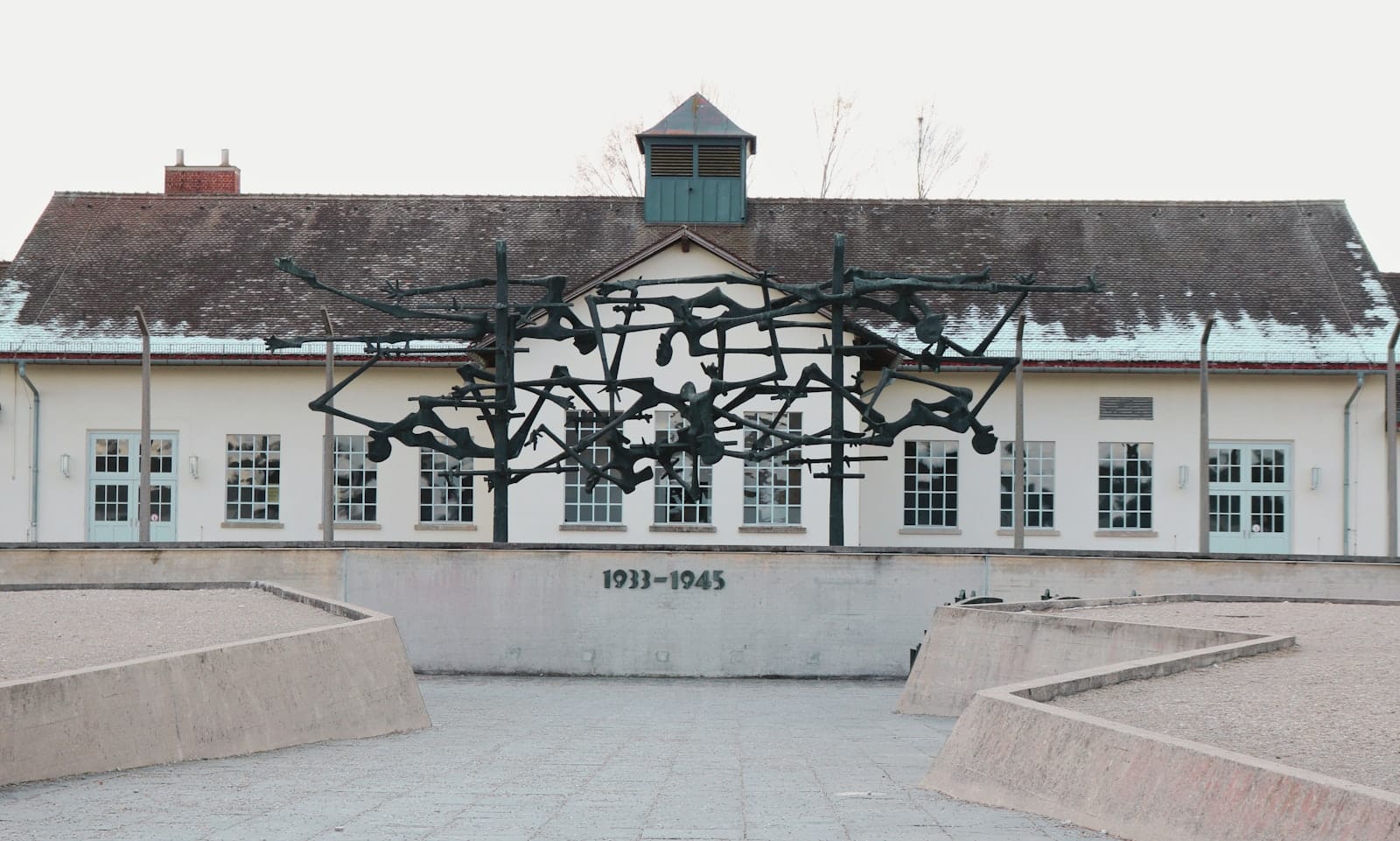 ด้านนอกของอนุสรณ์สถานค่ายกักกัน Dachau พร้อมประติมากรรมเหล็กที่โดดเด่นและจารึก 1933-1945 ที่มองเห็นได้ด้านหน้า