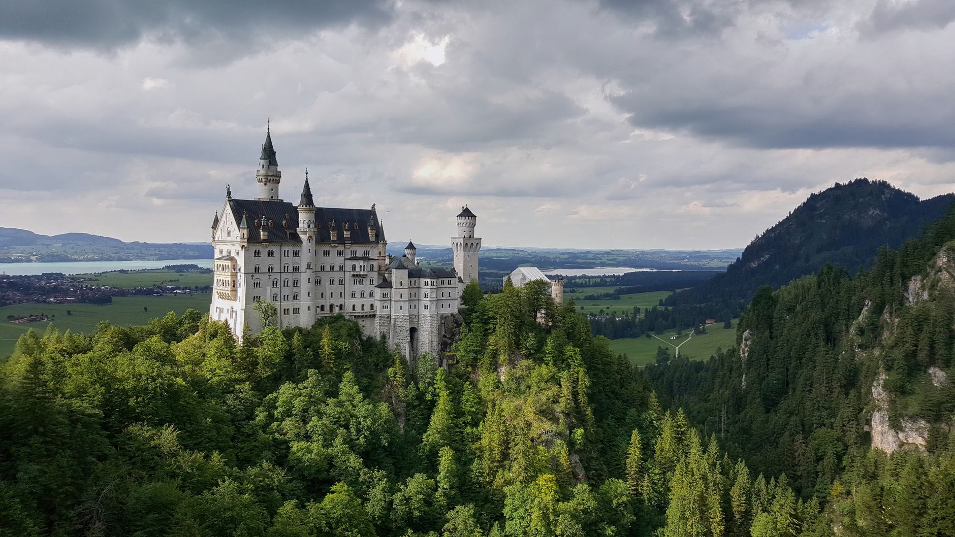 ปราสาท Neuschwanstein ตั้งอยู่บนเนินเขาที่ปกคลุมด้วยป่าไม้ ล้อมรอบด้วยทุ่งหญ้าสีเขียว มีเมฆครึ้มเหนือปราสาท และภูเขาในระยะไกลเป็นฉากหลัง