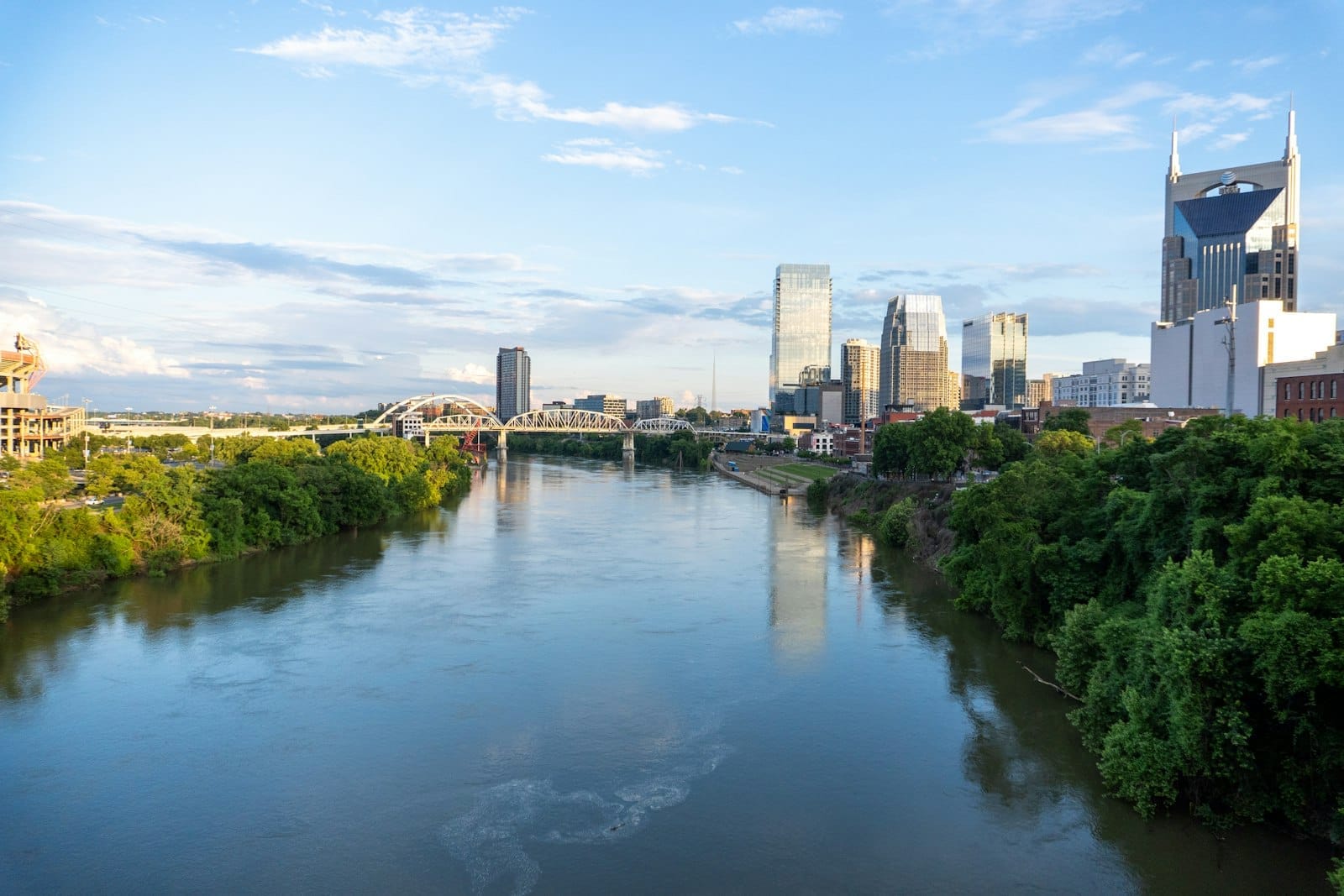Vista panorámica del horizonte de Nashville con un río en primer plano, árboles verdes a orillas del agua bajo un cielo azul intenso con nubes suaves.
