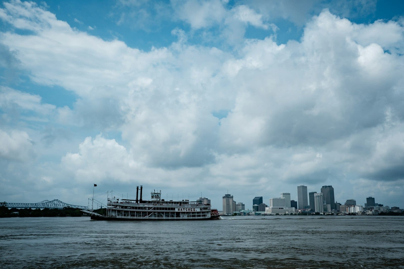 Um barco a vapor navega pelo rio Mississippi com o horizonte de Nova Orleans ao fundo sob um céu dramático com nuvens.