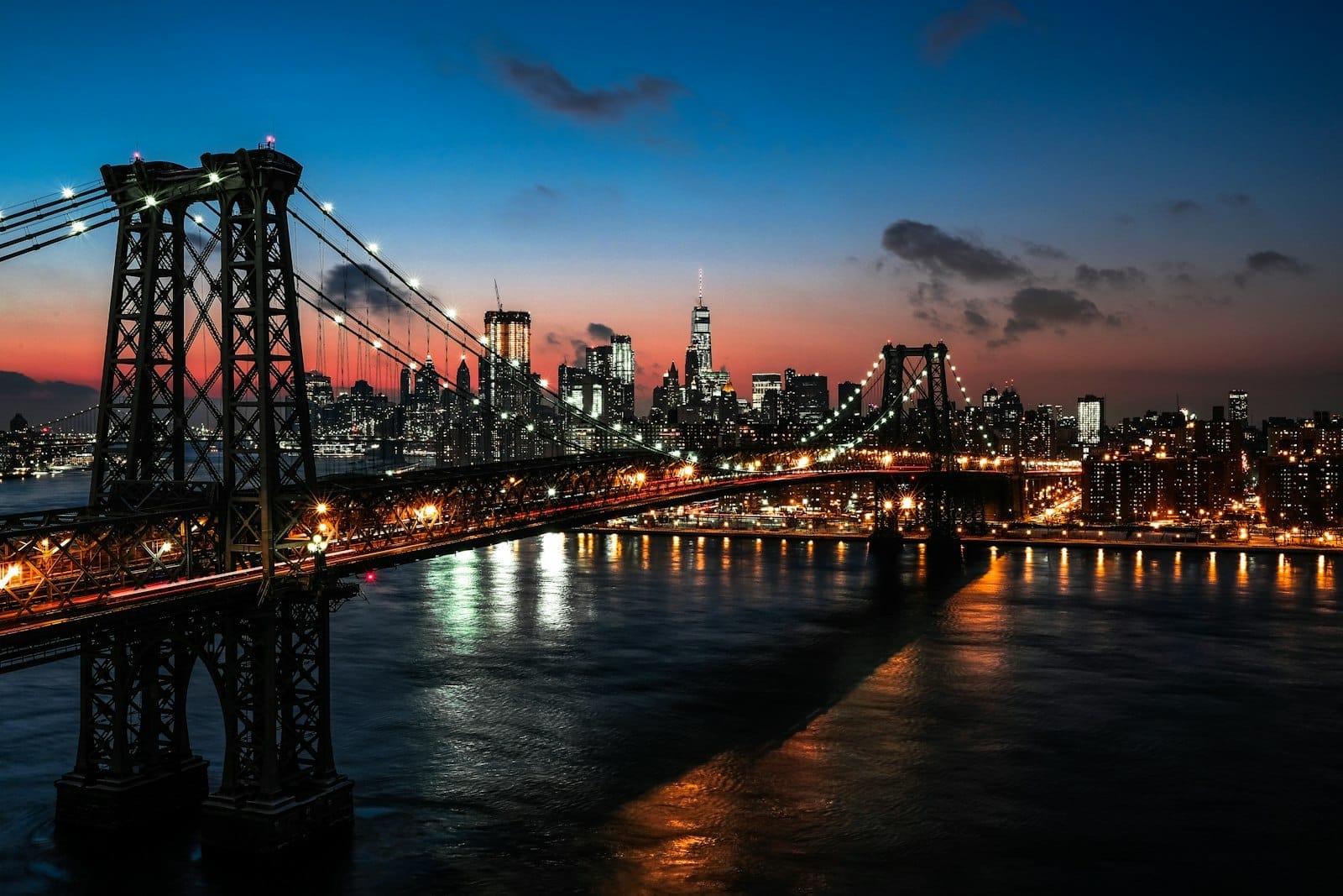 Vista impressionante de uma ponte iluminada ao pôr do sol com o skyline de Nova York ao fundo e o reflexo das luzes no rio abaixo.