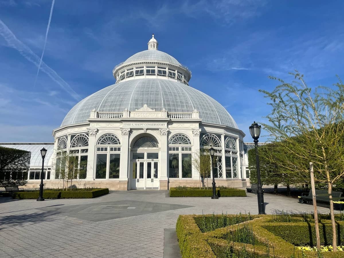 Das Glasgewächshaus mit Kuppeldach des New York Botanical Garden an einem klaren, sonnigen Tag, umgeben von gepflegten Gärten und Wegen.