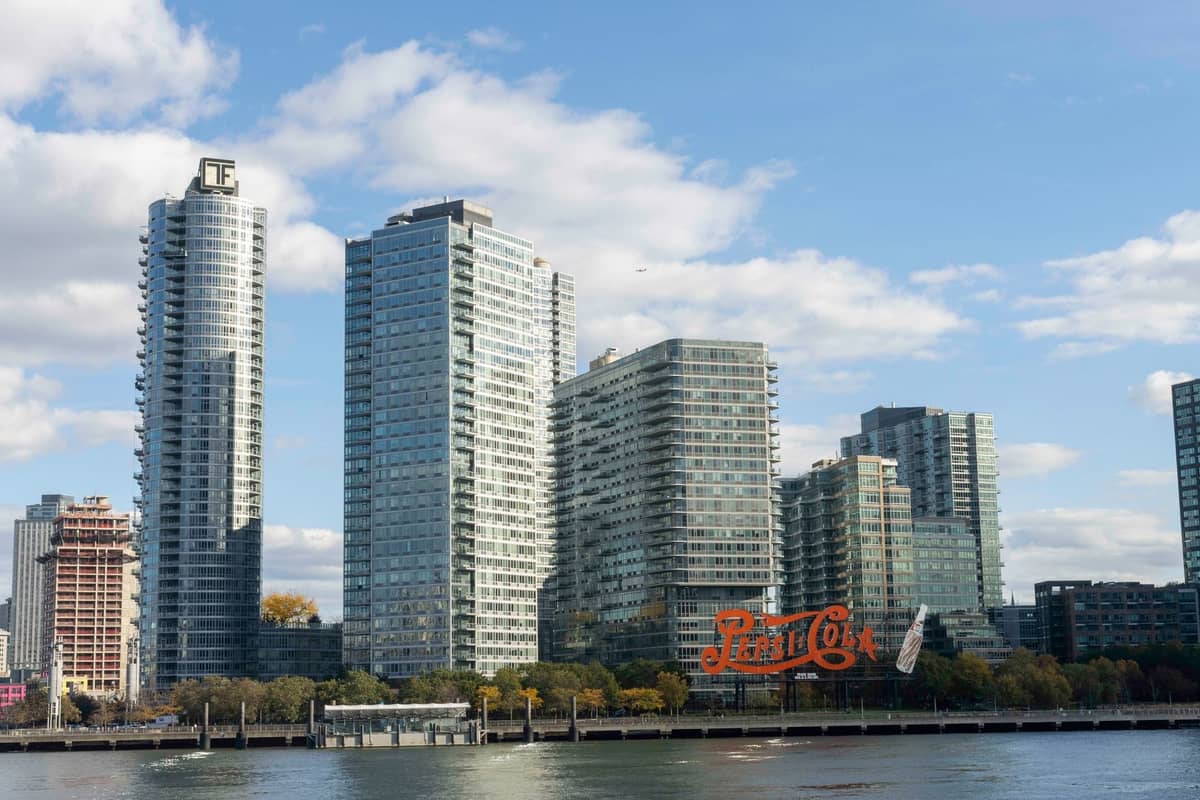 Das Ufer von Long Island City in Queens mit modernen Hochhäusern und dem ikonischen Pepsi-Cola-Schriftzug, von der anderen Seite des East River an einem sonnigen Tag fotografiert.