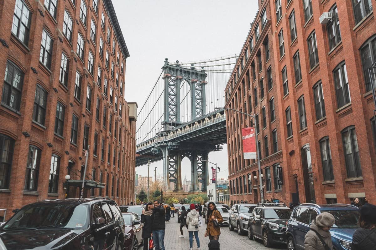 Menschen, die in DUMBO Brooklyn eine Straße entlanggehen, mit der Manhattan Bridge eingerahmt zwischen roten Backsteingebäuden.